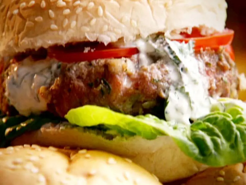 Spiced lamb burgers is topped with herbed yoghurt, tomatoes, and served with lettuce on a hamburger bun with sesame seeds.