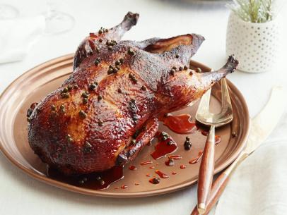 A close up of a roasted duck that is getting a glaze poured on it. The glaze is made from champagne vinegar, green pepper corns, soy sauce, honey and a dry marsala wine. The glaze is thick and running over the duck. The duck is sitting on a dark brown wood surface.