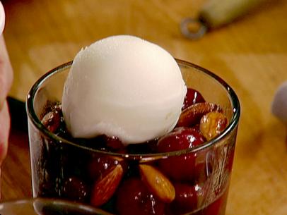 A view of sauteed cherries with grappa and almonds. The dessert has sorbet on top of it. The cherries are dark red and pitted. The almonds have been coated in butter and roasted in a pan. The dessert sits in a clear glass dessert dish. The dish is sitting on a dark brown wood surface.