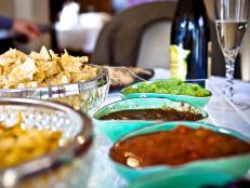 Table of salsa, guacamole, bean dip, and corn chips. Party setting with good wine and champagne.