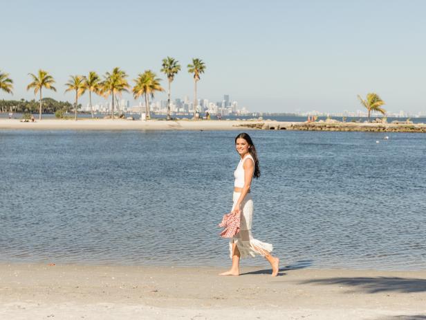 Host Katie Lee walks along the shore of the Redfish Grill restaurant grounds, with Miami's city scape in the background, as seen on Cooking Channel's Beach Bites with Katie Lee, Season 1.