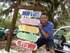 Host Sabin Lomac Posing Next to the hanging signage out front of Lucy Bell's in Daufuskie Island, South Carolina as seen on Cooking Channel's Seaside Snacks and Shacks, Season 1.